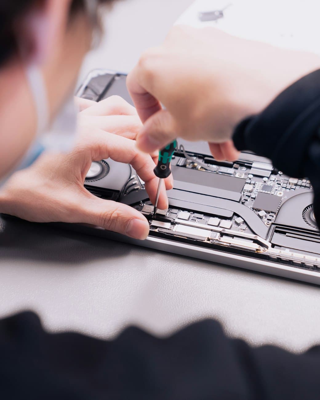 A BHCR technician at the bench, mid-repair on a laptop's logic board with a precision screwdriver